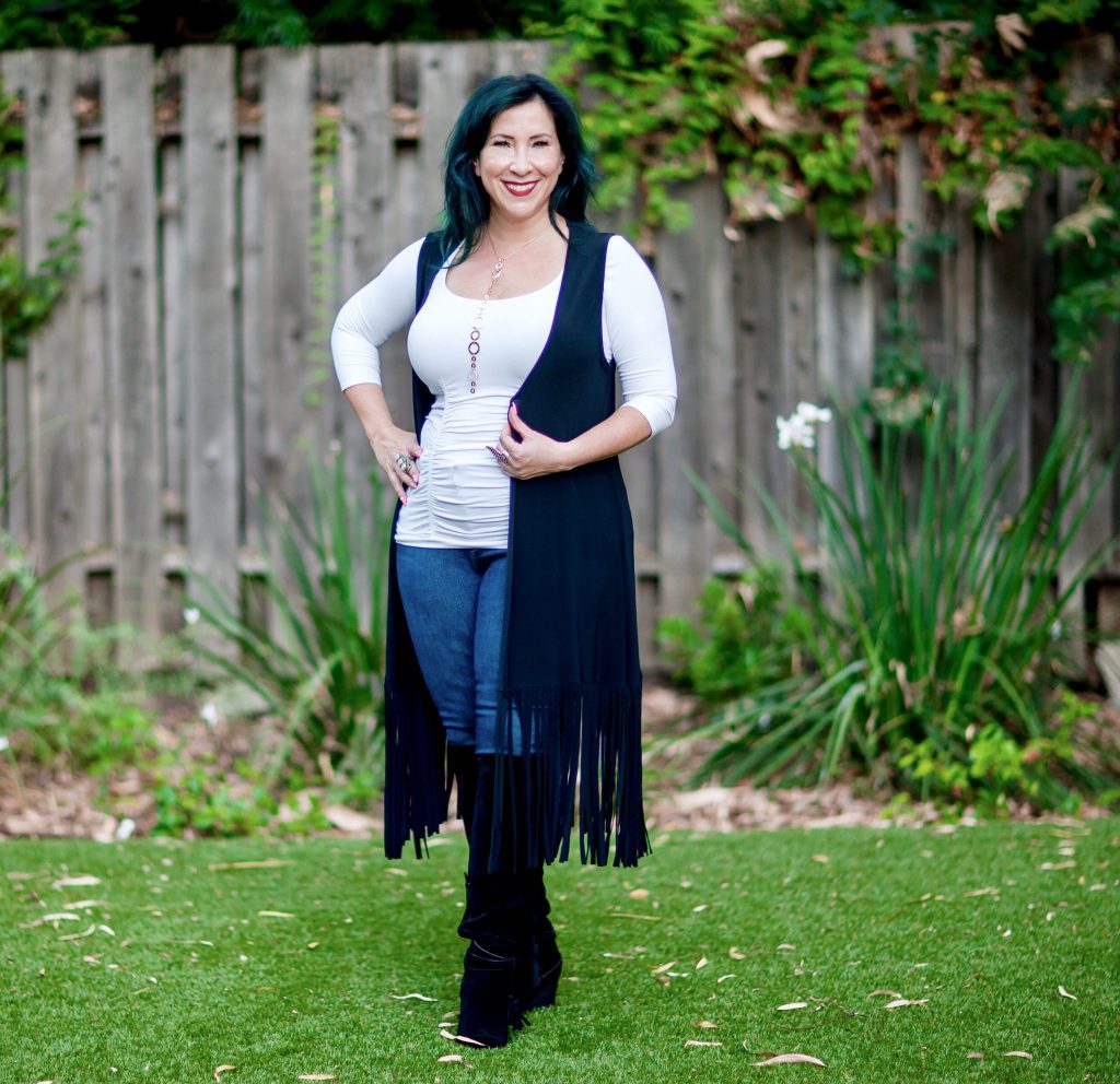 Woman in a white long sleeve top with a fringed vest, jeans and boots. 