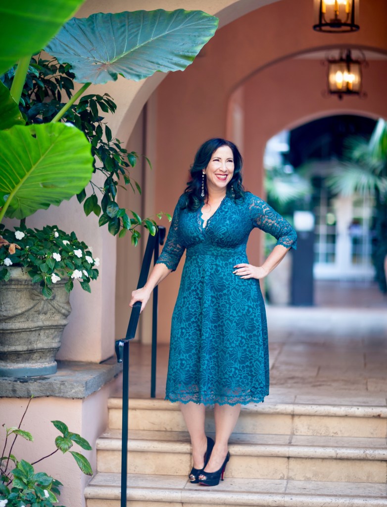 Woman in a turquoise Kiyonna dress made of lace standing on some steps.